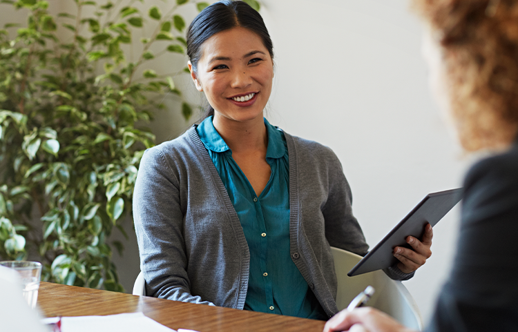 A financial advisor smiles and sits with her client while holding a tablet.